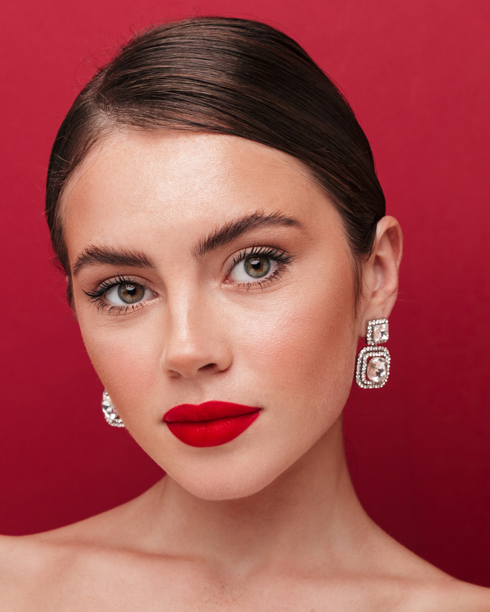 Glamorous holiday beauty portrait featuring a model with pigmented matte red lipstick, dewy skin, sleek brunette hair, and crystal statement earrings against a vibrant red backdrop